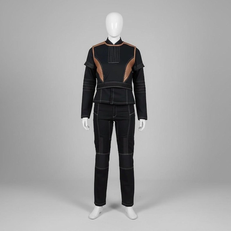 Op de afbeelding: Een zwart tweedelig outfit op een mannequin. De top heeft een hoge kraag en koperkleurige accenten op de schouders en borst. De broek is zwart met witte stiksels. De outfit is een futuristisch ontwerp.