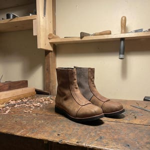 May include: A pair of brown leather ankle boots with a side zipper. The boots have a rounded toe and a dark sole. The boots are displayed on a wooden surface.