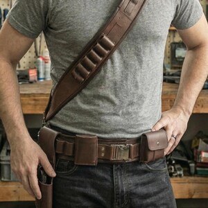 K&ouml;nnte beinhalten: Eine wei&szlig;e Keramik-Tasse mit schwarzem Henkel und dem Text "But First, Coffee" in fetter, schwarzer Schrift. Die Tasse steht vor einem verschwommenen Hintergrund, der eine gem&uuml;tliche Umgebung suggeriert.