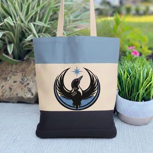 May include: A canvas tote bag with a three-tone design in light blue, beige, and black. The bag features a graphic of a bird with outstretched wings and a star. The handles are beige.