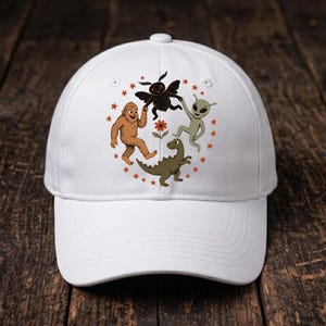 May include: A white baseball cap featuring a circular graphic print of a brown Bigfoot, a black moth-like creature, a green alien, and a green dinosaur surrounding a small orange flower, all encircled by small orange stars on the front panel.