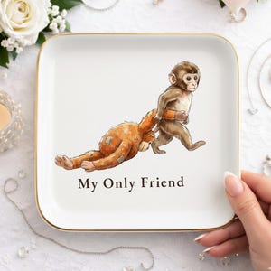 May include: A square, white trinket tray with gold trim. The tray features a whimsical illustration of two monkeys, one pulling the other. The text "My Only Friend" is printed below the monkeys.
