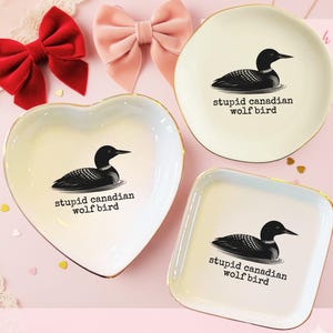 May include: Three decorative ceramic dishes in heart, round, and square shapes. Each dish is cream-colored with a gold rim and features a black and white illustration of a loon and the text "stupid canadian wolf bird."