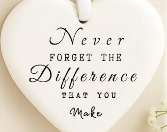 May include: A white heart-shaped ornament with the text "Never Forget the Difference That You Make" in black script. The ornament hangs from a white ribbon and rests on a cream-colored box.