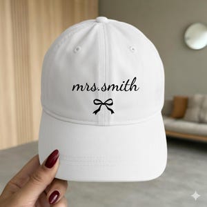 May include: A white baseball cap featuring the black text "mrs.smith" in a cursive font, positioned above a small black graphic of a tied bow. The cap has a structured crown, a curved brim, and eyelets for ventilation on the top panels.
