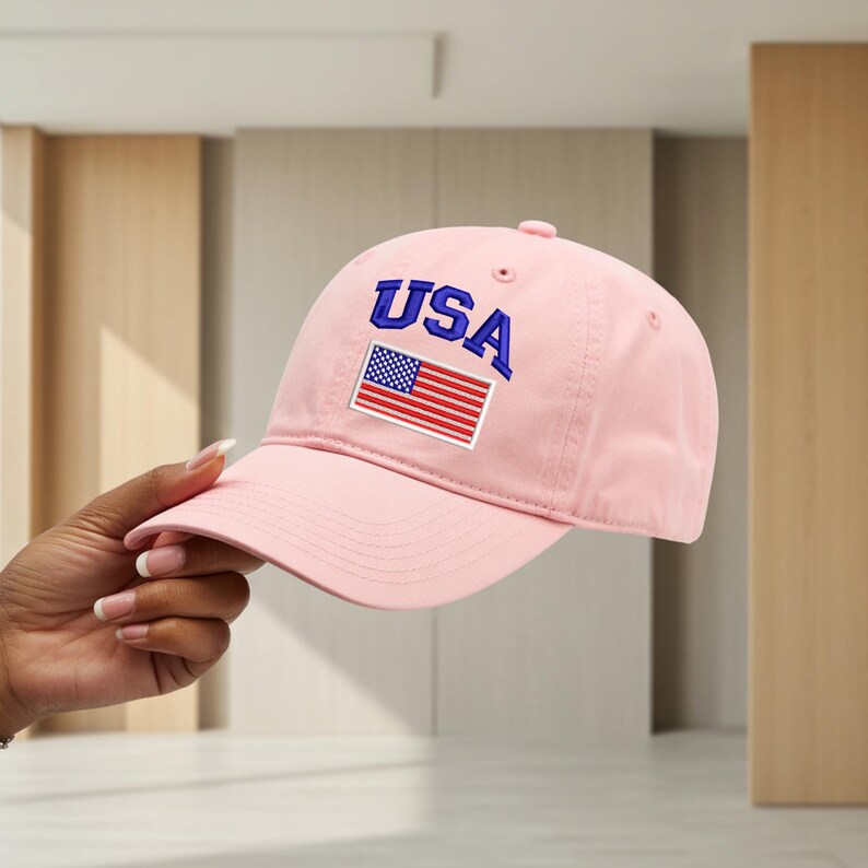 May include: A light pink baseball cap featuring the text USA in bold blue letters above a rectangular patch of the American flag. The cap has a curved brim, a button on the top, and stitched eyelets for ventilation on the crown panels.