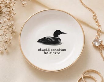 May include: A round white ceramic trinket dish with a gold-colored rim. The center features a black graphic illustration of a loon bird above the black typewriter-style text that reads, "stupid canadian wolf bird" in a clean, centered layout.