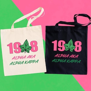 May include: Two tote bags, one cream and one black, with the text "1948" in pink, a green ivy leaf, and the words "ALPHA AKA ALPHA KAPPA" in green. Both bags have handles.