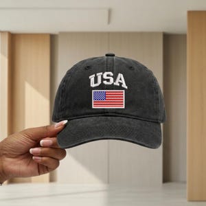 May include: A charcoal grey baseball cap with a distressed, washed-out finish. The front features the white embroidered text USA arched above a rectangular patch displaying the American flag with its traditional red, white, and blue colors.
