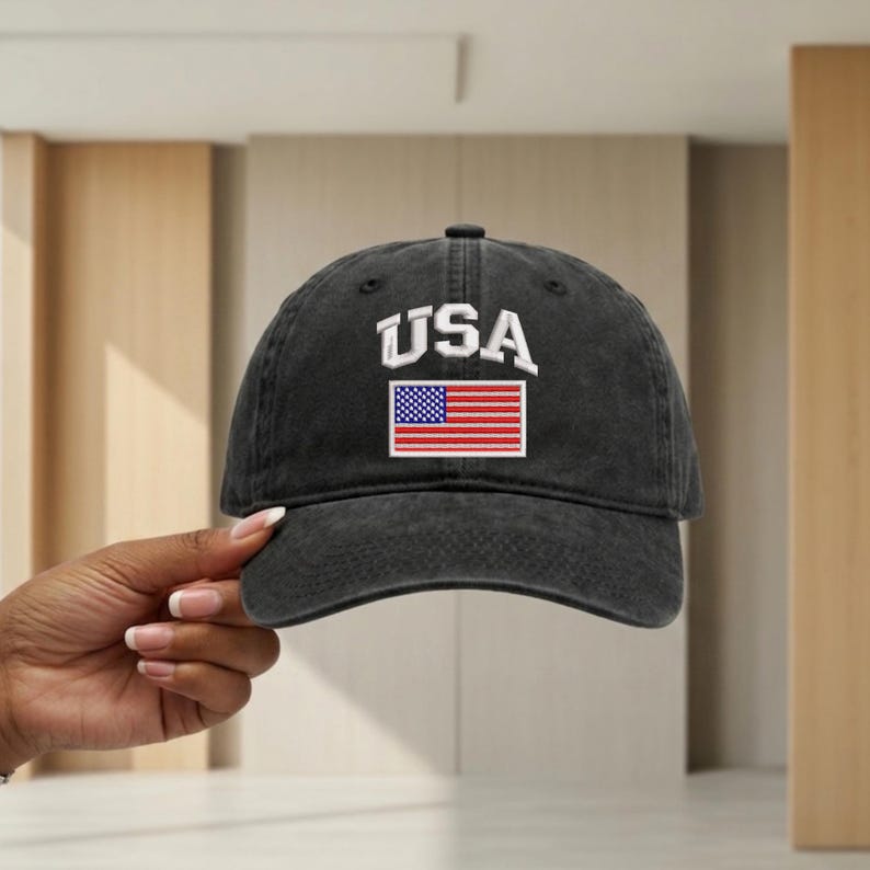 May include: A charcoal grey baseball cap with a distressed, washed-out finish. The front features the white embroidered text USA arched above a rectangular patch displaying the American flag with its traditional red, white, and blue colors.