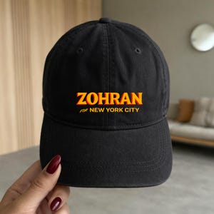 May include: Black baseball cap with the word "ZOHRAN" in yellow and orange block letters, with "FOR NEW YORK CITY" below. The cap has a curved brim and a rounded top.