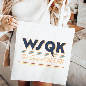 May include: White tote bag with the text "WSQK The Squawk 94.5 FM" in navy blue and orange lettering. The design includes horizontal lines in teal, yellow, and red. The bag has cream-colored handles.