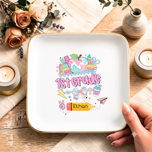 May include: A square white ceramic trinket dish with a gold rim featuring a colorful graphic of school supplies, a floral apple, a bow, and the text 1st Grade. A yellow pencil graphic at the bottom includes the personalized name Ethan.