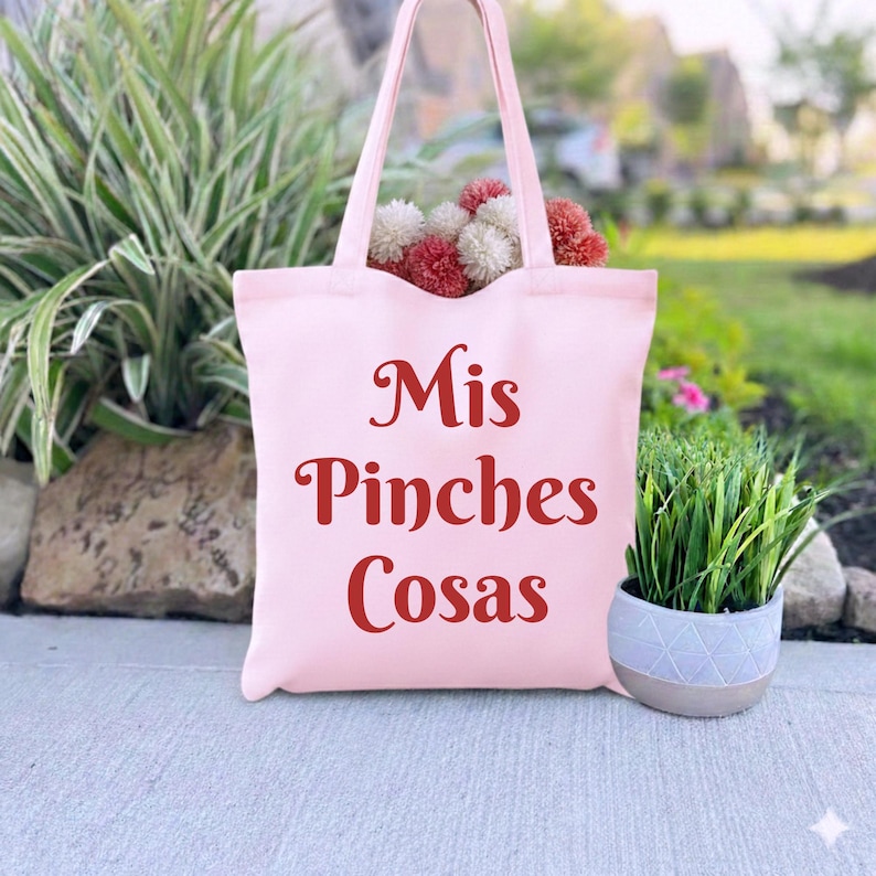 K&ouml;nnte beinhalten: Eine hellrosa Tragetasche mit dem Text "Mis Pinches Cosas" in burgunderfarbener Schrift. Die Tasche ist mit dekorativen Blumen gef&uuml;llt. Rechts neben der Tasche steht eine kleine Topfpflanze.