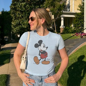 May include: Light blue t-shirt featuring a graphic of Mickey Mouse in black, red, and brown. The shirt has short sleeves and a crew neck. The Disney logo is printed below the graphic.