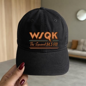 May include: Black baseball cap with the text "WSQK The Squawk 94.5 FM" in orange and blue. The cap has a curved brim and a rounded crown. The hat is made of a soft, durable fabric.