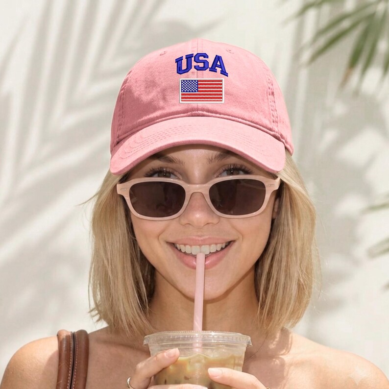 May include: A light pink baseball cap featuring the text USA in blue embroidery above a rectangular patch displaying the American flag. The cap has a curved brim and a structured front panel, providing a classic look with a vibrant color contrast.