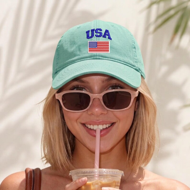 May include: A light mint green baseball cap featuring the text USA embroidered in blue above a rectangular patch of the American flag. The cap has a curved brim and a structured crown with visible stitching details across the front panels.