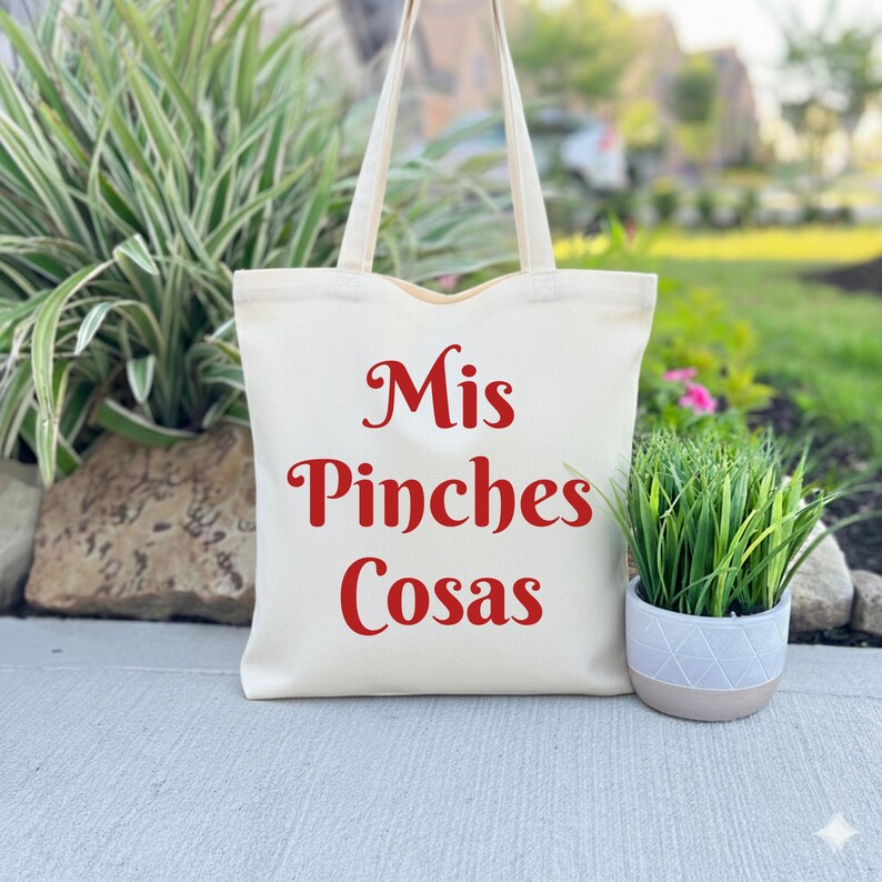 K&ouml;nnte beinhalten: Cremefarbene Tragetasche mit rotem Text "Mis Pinches Cosas". Die Tasche hat zwei Griffe und steht auf einer Betonoberfl&auml;che neben einer kleinen Topfpflanze mit gr&uuml;nem Gras. Der Hintergrund umfasst Gr&uuml;n und ein Haus.
