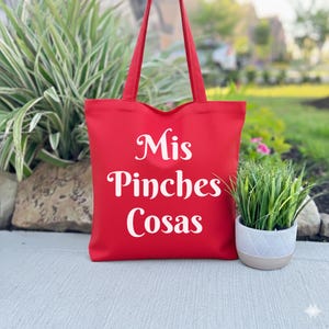 K&ouml;nnte beinhalten: Eine rote Tragetasche mit wei&szlig;em Text "Mis Pinches Cosas". Die Tasche hat zwei rote Griffe und steht auf einer Betonoberfl&auml;che. Rechts neben der Tasche befindet sich eine kleine Topfpflanze.