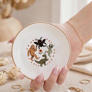May include: A round white ceramic trinket dish with a gold-toned rim. The center features a colorful illustration of a brown Bigfoot, a black moth-like creature, a green alien, and a green dinosaur holding hands around a small orange flower.
