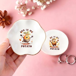 May include: Two white ceramic trinket dishes with gold-toned rims. The larger dish features a cartoon sweet potato character and the text "SHE IS MY sweet POTATO". The smaller dish features a cartoon potato character and the text "HE IS MY sweet POTATO".