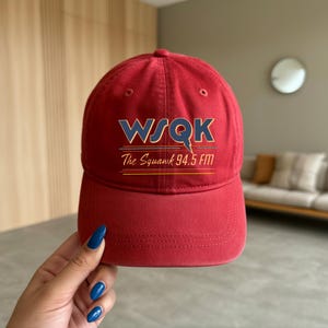May include: A red baseball cap with the text "WSQK The Squawk 94.5 FM" in blue and gold. The cap has a curved brim and a rounded crown. The hat is made of a soft material.