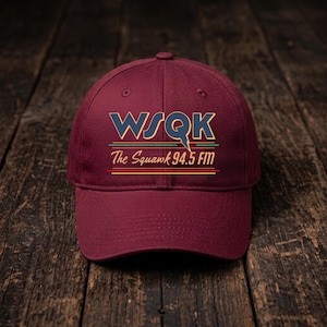 May include: A maroon baseball cap featuring a graphic print with the text WSQK and The Squawk 94.5 FM in blue, yellow, and teal lettering. The design includes horizontal lines and a lightning bolt detail integrated into the letter Q.