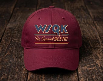 May include: A maroon baseball cap featuring a graphic print with the text WSQK and The Squawk 94.5 FM in blue, yellow, and teal lettering. The design includes horizontal lines and a lightning bolt detail integrated into the letter Q.