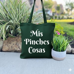 K&ouml;nnte beinhalten: Eine dunkelgr&uuml;ne Tragetasche mit wei&szlig;em Text "Mis Pinches Cosas". Die Tasche hat lange Griffe und ist aus robustem Material gefertigt. Die Tragetasche steht auf einer Betonoberfl&auml;che neben einer kleinen Topfpflanze.