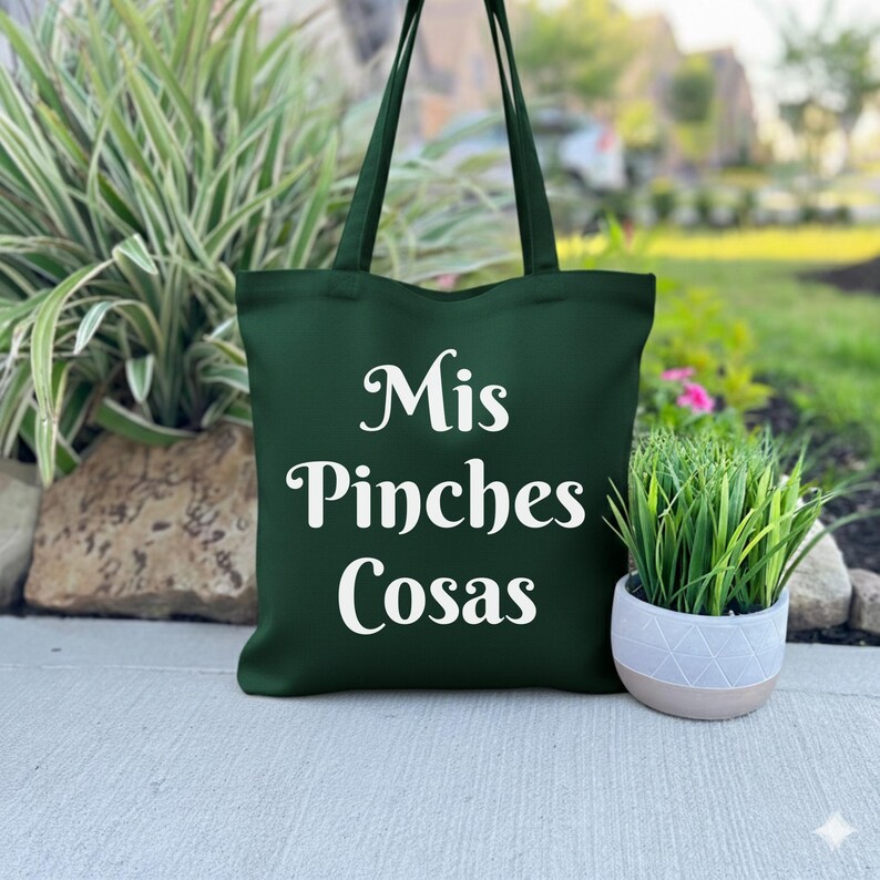 K&ouml;nnte beinhalten: Eine dunkelgr&uuml;ne Tragetasche mit wei&szlig;em Text "Mis Pinches Cosas". Die Tasche hat lange Griffe und ist aus robustem Material gefertigt. Die Tragetasche steht auf einer Betonoberfl&auml;che neben einer kleinen Topfpflanze.