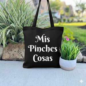 K&ouml;nnte beinhalten: Schwarze Tragetasche mit dem Text "Mis Pinches Cosas" in wei&szlig;er Schrift. Die Tasche hat lange Henkel und steht auf einer Betonoberfl&auml;che neben einer kleinen Topfpflanze. Der Hintergrund umfasst Gr&uuml;n und eine verschwommene Ansicht eines Gartens.