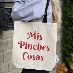 K&ouml;nnte beinhalten: Cremefarbene Tragetasche mit dem roten Schriftzug "Mis Pinches Cosas". Die Tasche hat schwarze Tr&auml;ger und wird von einer Person in einem grauen Sweatshirt gehalten. Im Hintergrund sind eine Backsteinmauer und Gr&uuml;n zu sehen.