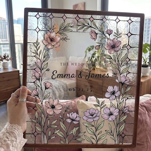 May include: A rectangular stained glass panel in a dark wood frame featuring a floral wreath design with pink flowers and green leaves. The center text reads THE WEDDING OF Emma & James 06.22.25. The glass has a textured, grid-like pattern.