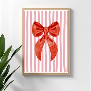 May include: A framed art print featuring a large, watercolor-style red bow against a background of pink and white vertical stripes. The artwork is in a light wood frame, suitable for wall decor.