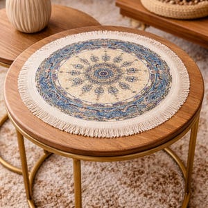 Puede incluir: Una mesa auxiliar redonda de madera con base de metal dorado. La mesa presenta un tapete decorativo circular con fondo crema y diseño floral azul y dorado. El tapete tiene un borde con flecos.