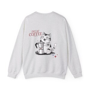 May include: Light grey sweatshirt with a cartoon cat and coffee maker design. The text "DECAF COFFEE" is above the cat. The sweatshirt is a casual style, suitable for everyday wear.