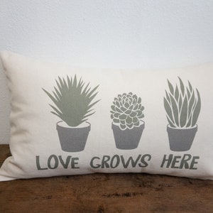 May include: A white pillow with a green silhouette of three potted plants and the text "LOVE GROWS HERE" printed on it.