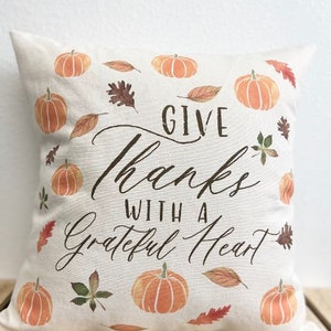 May include: A white throw pillow with the text "Give Thanks With A Grateful Heart" in brown cursive lettering. The pillow is decorated with orange pumpkins and brown leaves.