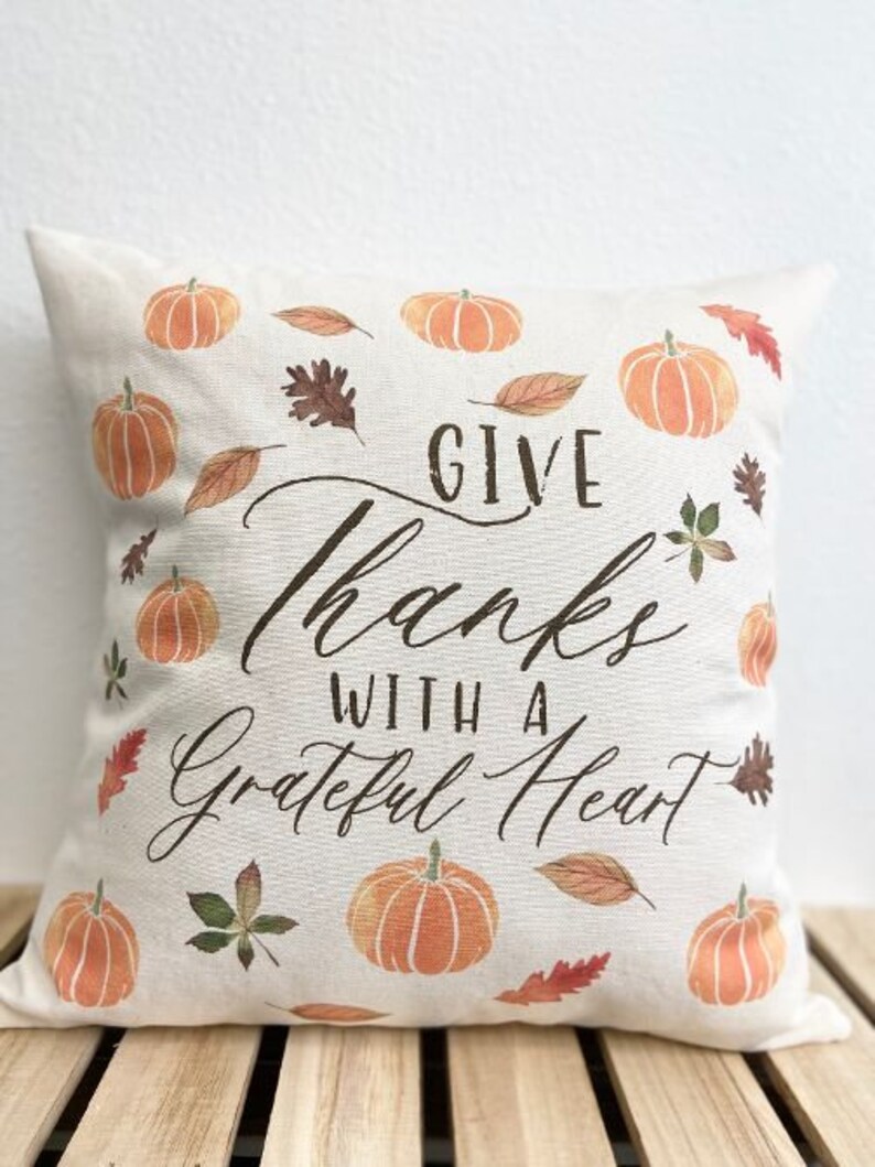 May include: A white throw pillow with the text "Give Thanks With A Grateful Heart" in brown cursive lettering. The pillow is decorated with orange pumpkins and brown leaves.