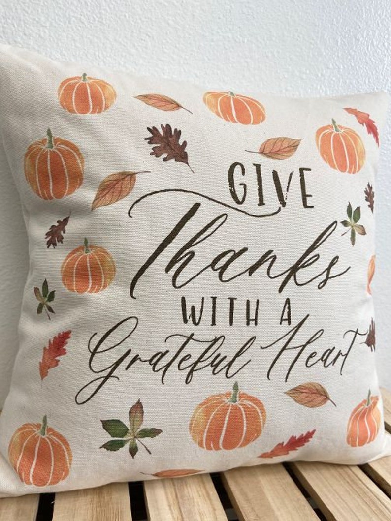 May include: A white throw pillow with a fall theme. It features a brown and orange pumpkin design, brown leaves, and the text "Give Thanks With A Grateful Heart".