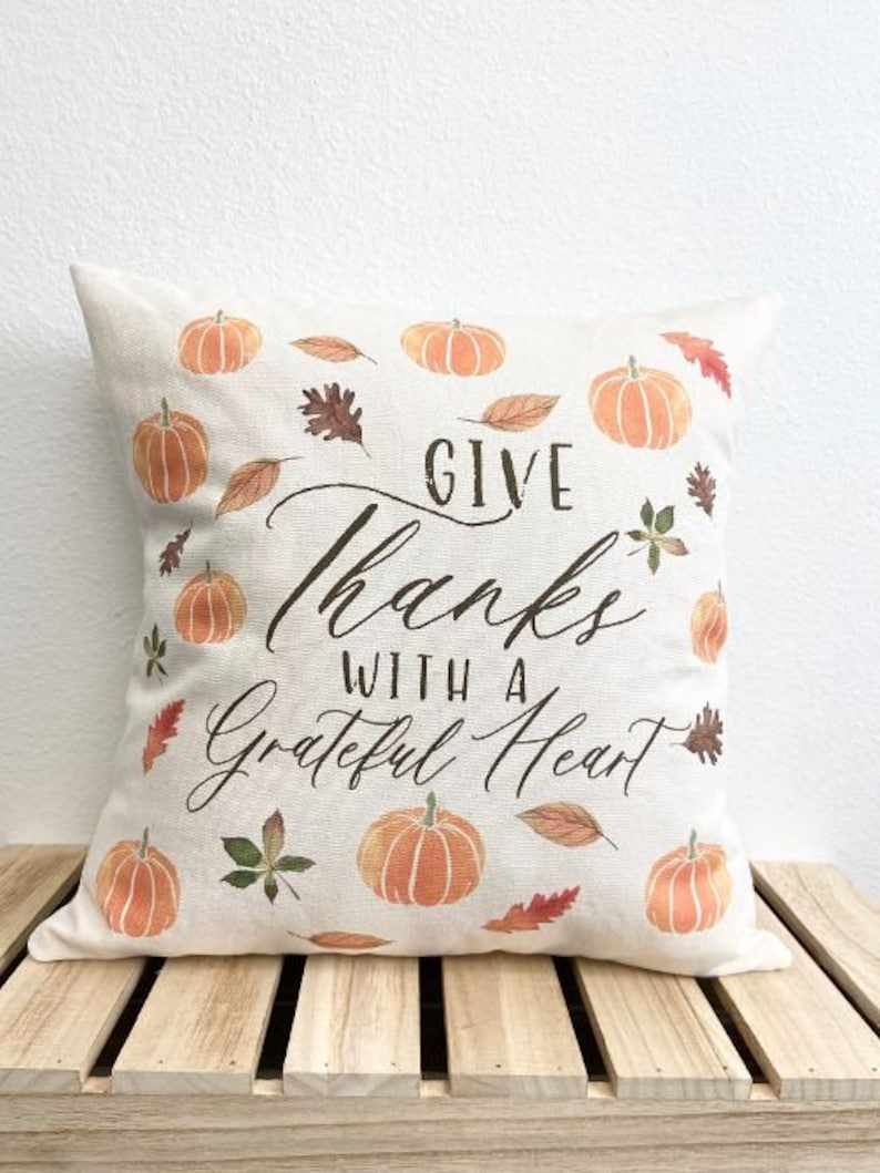 May include: A white throw pillow with a brown and white design featuring pumpkins and leaves. The pillow has the text "Give Thanks With A Grateful Heart" printed on it.