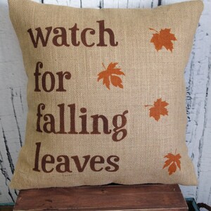 May include: A brown burlap pillow with the text "watch for falling leaves" and orange leaf silhouettes printed on it.