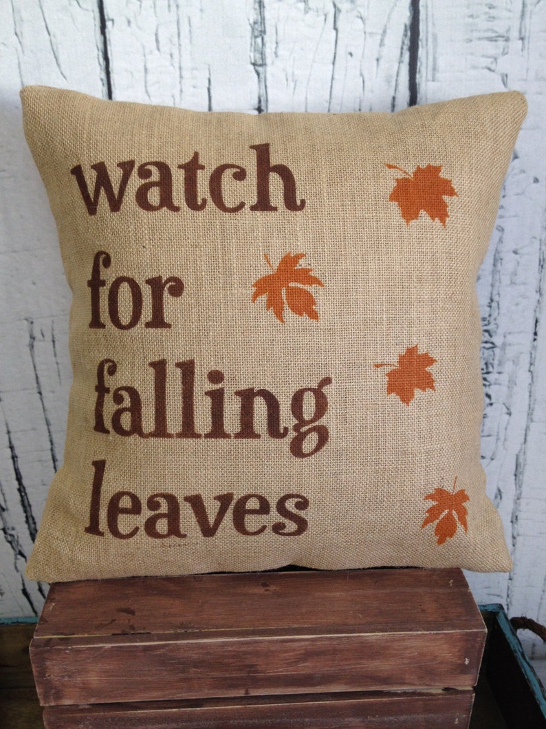 May include: A brown burlap pillow with the text "watch for falling leaves" and orange leaf silhouettes printed on it.