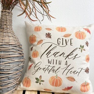 May include: A beige throw pillow with the text "Give Thanks With A Grateful Heart" in black script. The pillow is decorated with orange pumpkins and brown leaves.