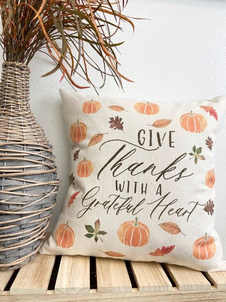 May include: A beige throw pillow with the text "Give Thanks With A Grateful Heart" in black script. The pillow is decorated with orange pumpkins and brown leaves.