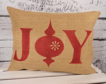 May include: A burlap pillow with the word "JOY" printed in red. The "O" is replaced with a red Christmas ornament with a white snowflake in the center.