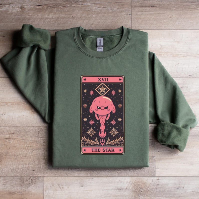 Puede incluir: Una sudadera verde oliva con un dise&ntilde;o de carta del tarot. La carta representa una criatura rosa con forma de hongo y cara enfadada, rodeada de estrellas y el texto "THE STAR" y "XVII".