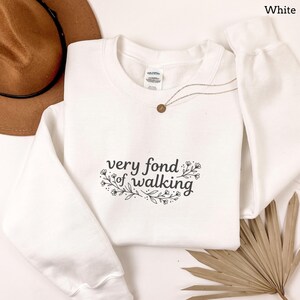 May include: White sweatshirt with the text "very fond of walking" in a black script font, surrounded by floral accents. A gold necklace with a small pendant is draped across the sweatshirt. A brown hat is visible in the background.