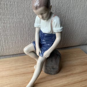 Può includere: Statuina in porcellana di un bambino seduto, con la testa china. Il bambino indossa una camicia bianca, pantaloncini blu e bretelle. La statuina è seduta su una pietra grigia. I capelli del bambino sono castani.